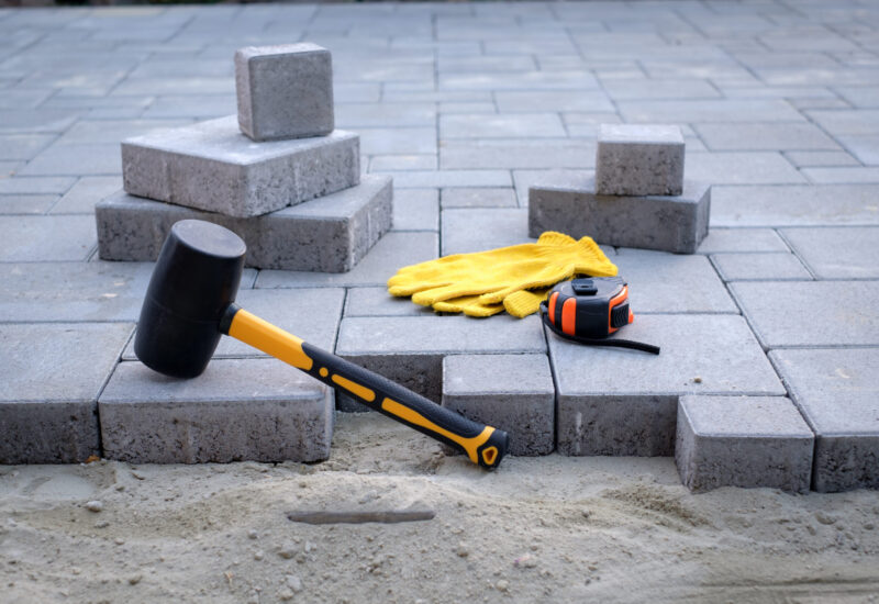 The master in yellow gloves lays paving stones in layers. Garden brick pathway paving by professional paver worker. Laying gray concrete paving slabs in house courtyard on sand foundation base.